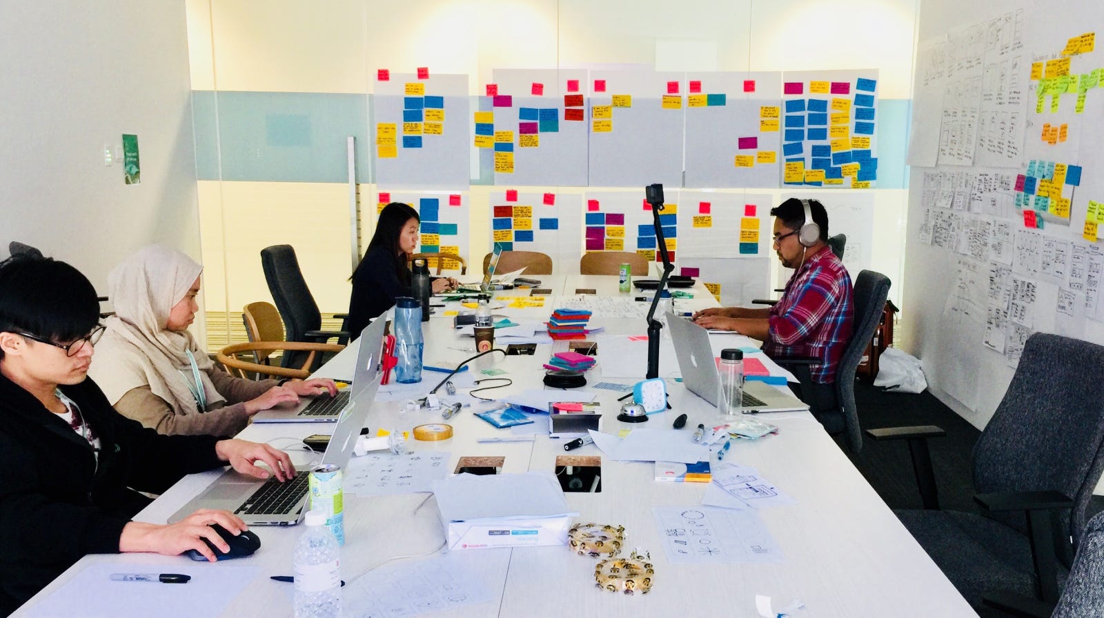 Four colleagues sitting around a large table surrounded by post-its and computers all working diligently on their laptops.