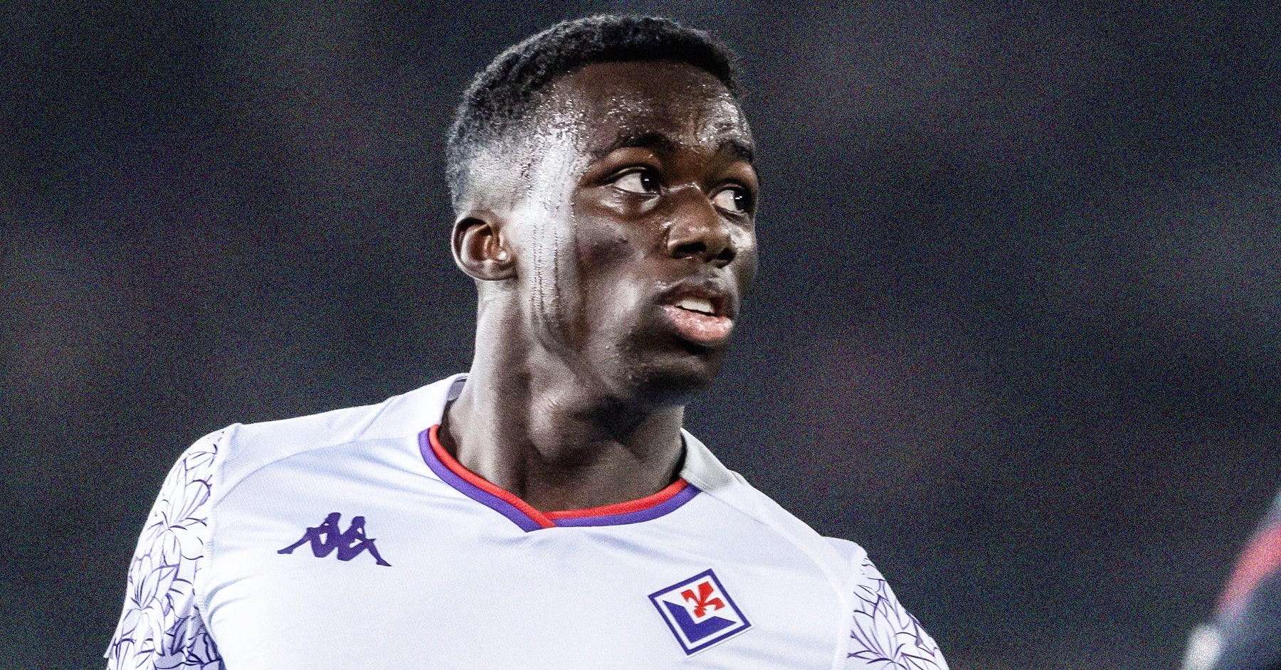 A photo of Michael Kayode wearing a white Fiorentina kit with purple trim set against a dark, blurred background.