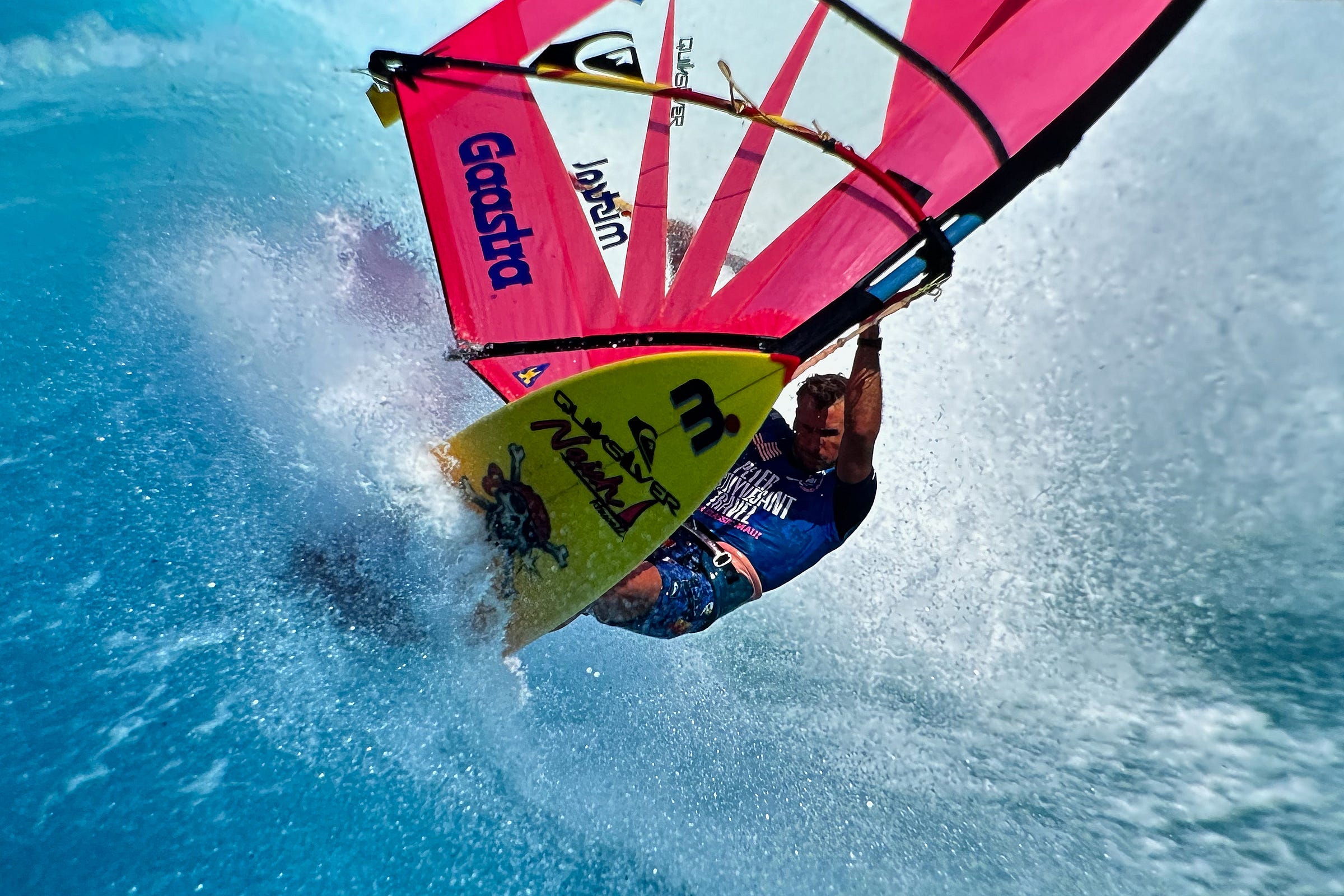 Close photograph of a windsurfer on a yellow surf board with a bright magenta sail on a large wave in Hawaii.