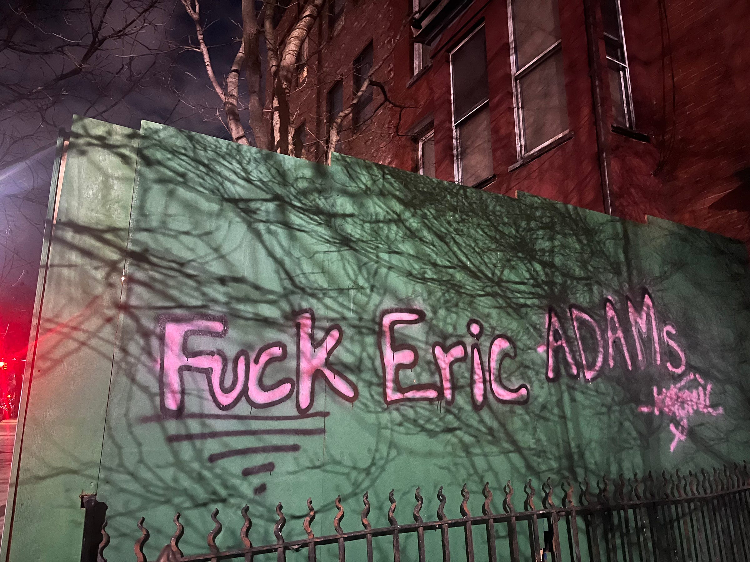At night, on some green temporary construction walling outside, are the words "FUCK ERIC ADAMS" spray painted in pink with black outlines