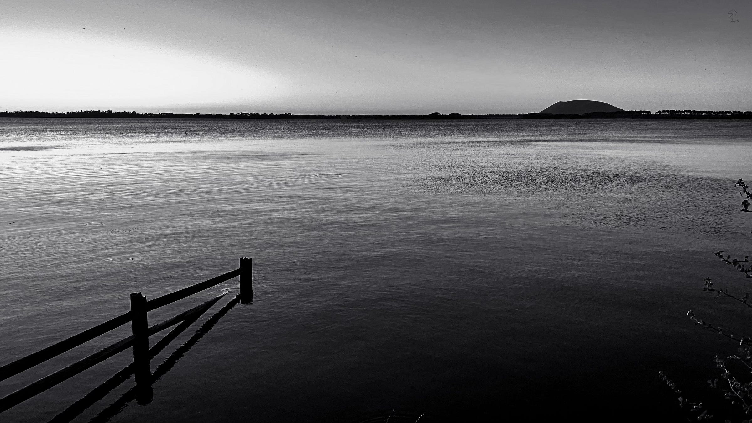 View at dusk across a large lake towards a steep-sided, flat-topped hill. The surface of the lake is lightly riffled by breeze. A half-sunken fence intrudes diagonally into the picture from bottom left