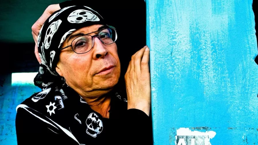 Pedro Lemembel usa lenço com estampa de caveiras, óculos e está encostado em uma parede azul clara