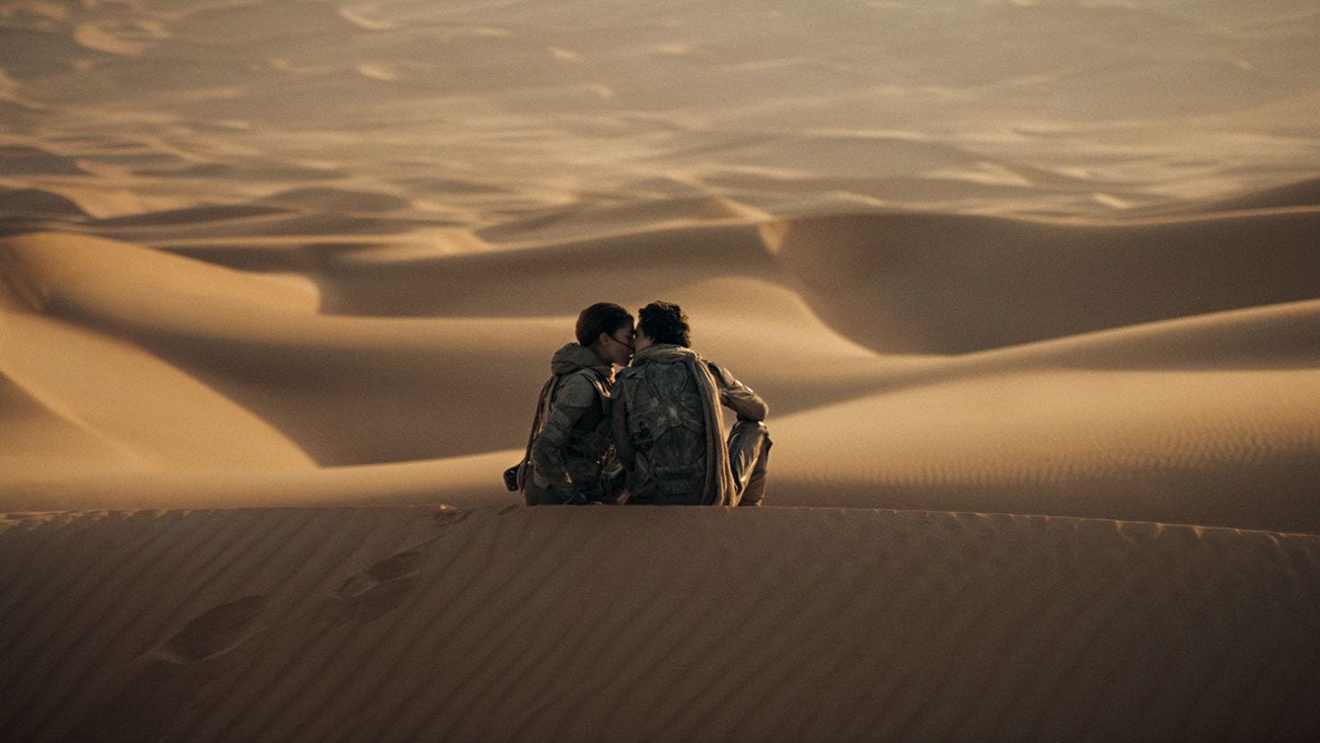 Actors Zendaya and Timothee Chalamet share a kiss while seated on a sand Dune in the movie Dune: Part Two. Both are dressed in gear from the Fremen people, and they are surrounded by sand in the rest of the frame.