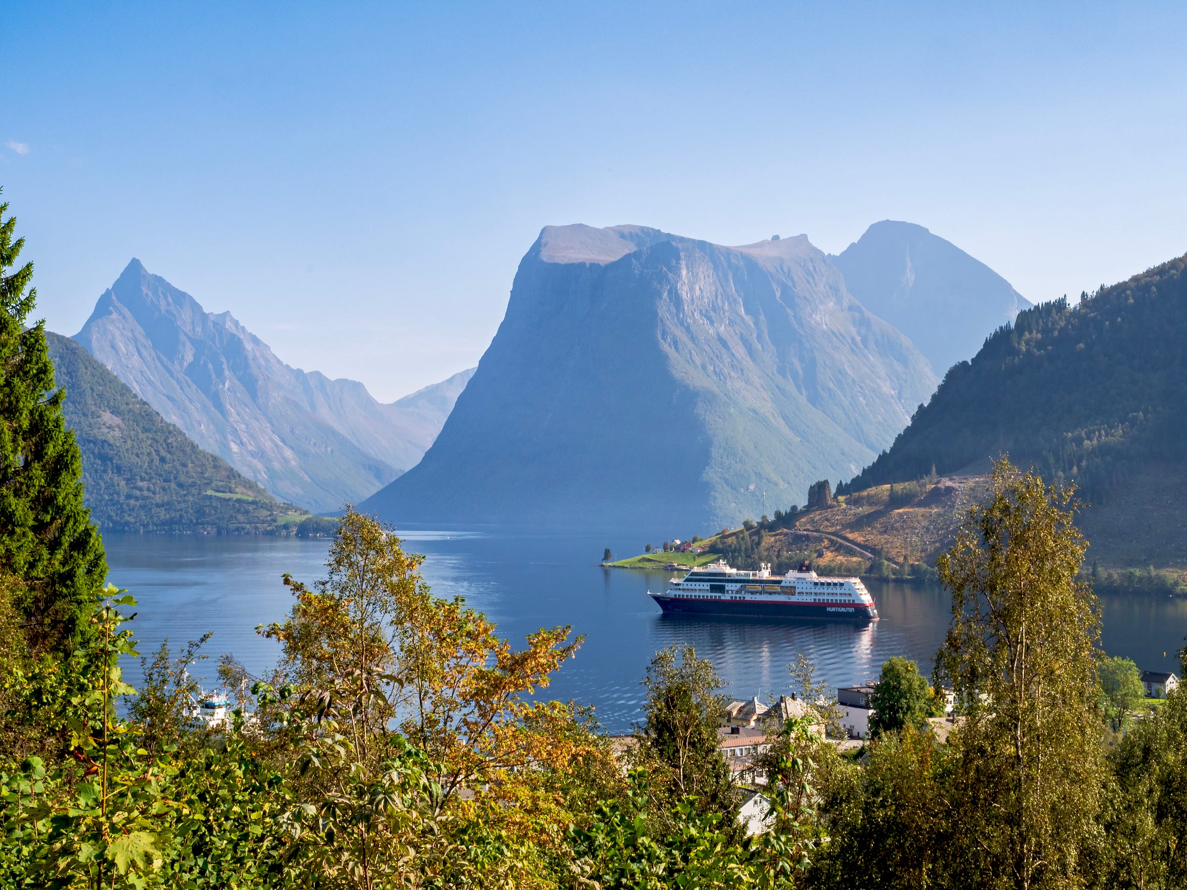 View over a Norwegian fjord to dramatic mountain peaks. There is vegetation in the foreground, and the rooftops of a village can just be seen.  A cruise ship is anchored in the fjord.