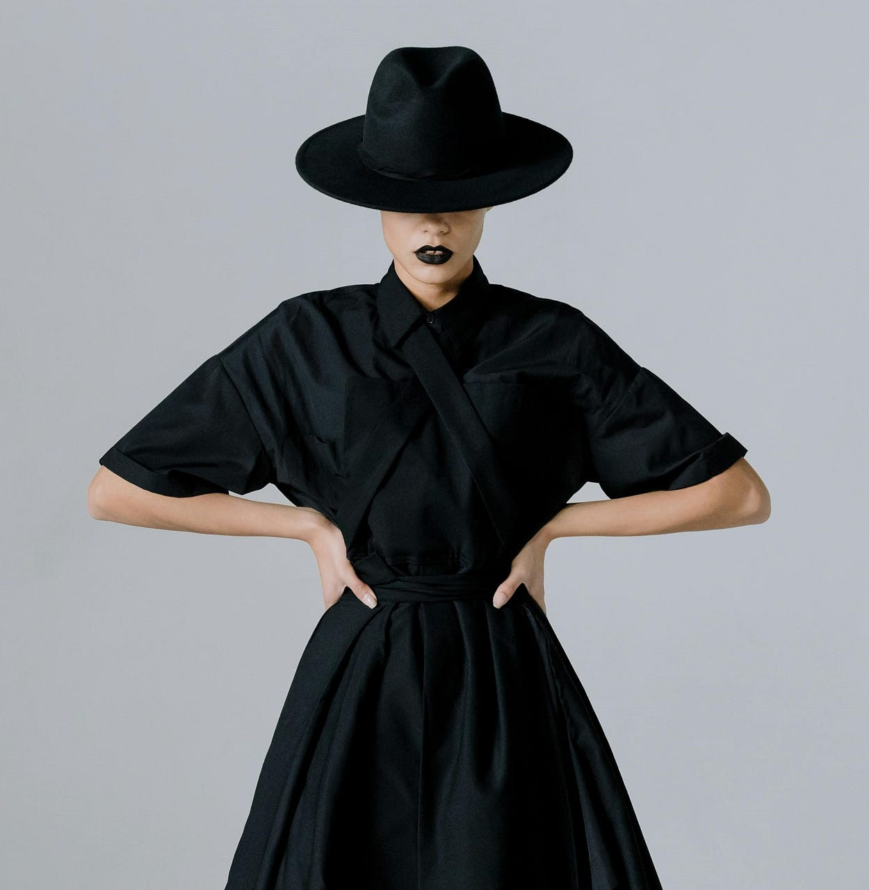 A woman poses with both hands on her waist. She’s wearing black lipstick, a black hat, and a black dress. The backdrop is gray. A woman poses with both hands on her waist. She’s wearing black lipstick, a black hat, and a black dress. The backdrop is gray.