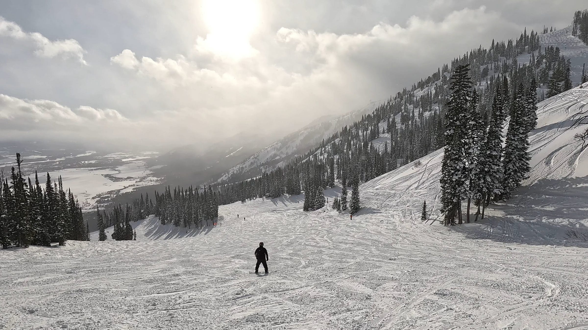 Photograph of a solitary snowboarder, clad in black against a white background of snow, mountains, and trees. Teton Village in Jackson Hole, WY can be seen in the distance at the foot of the mountain