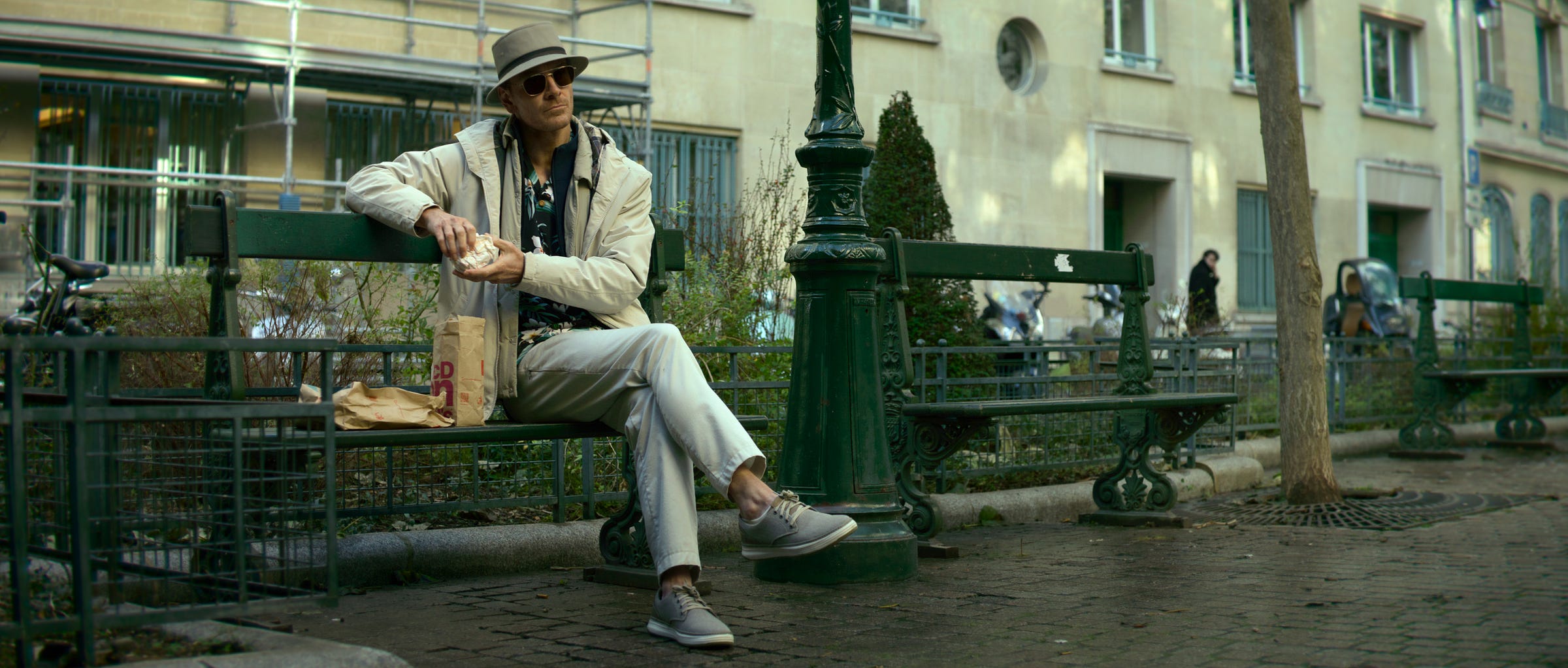 Michael Fassbender as The Killer sitting on a bench unwrapping a McDonald's sandwich