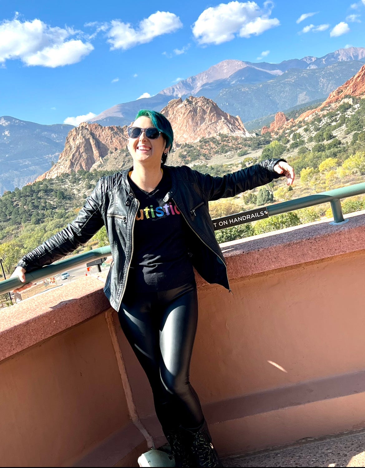 Lyric appears in all black. Leather jacket, leather pants, and tshirt with the word Autistic in a rainbow font. They are on a balcony, overlooking the Garden of the Gods Park