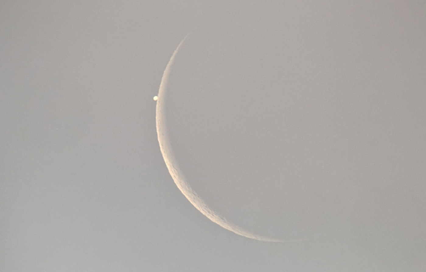 Jupiter is a small bright white disk just touching the edge of the Moon, which is far larger and a very thin crescent. A few lunar craters are visible. Jupiter is a small bright white disk just touching the edge of the Moon, which is far larger and a very thin crescent. A few lunar craters are visible.