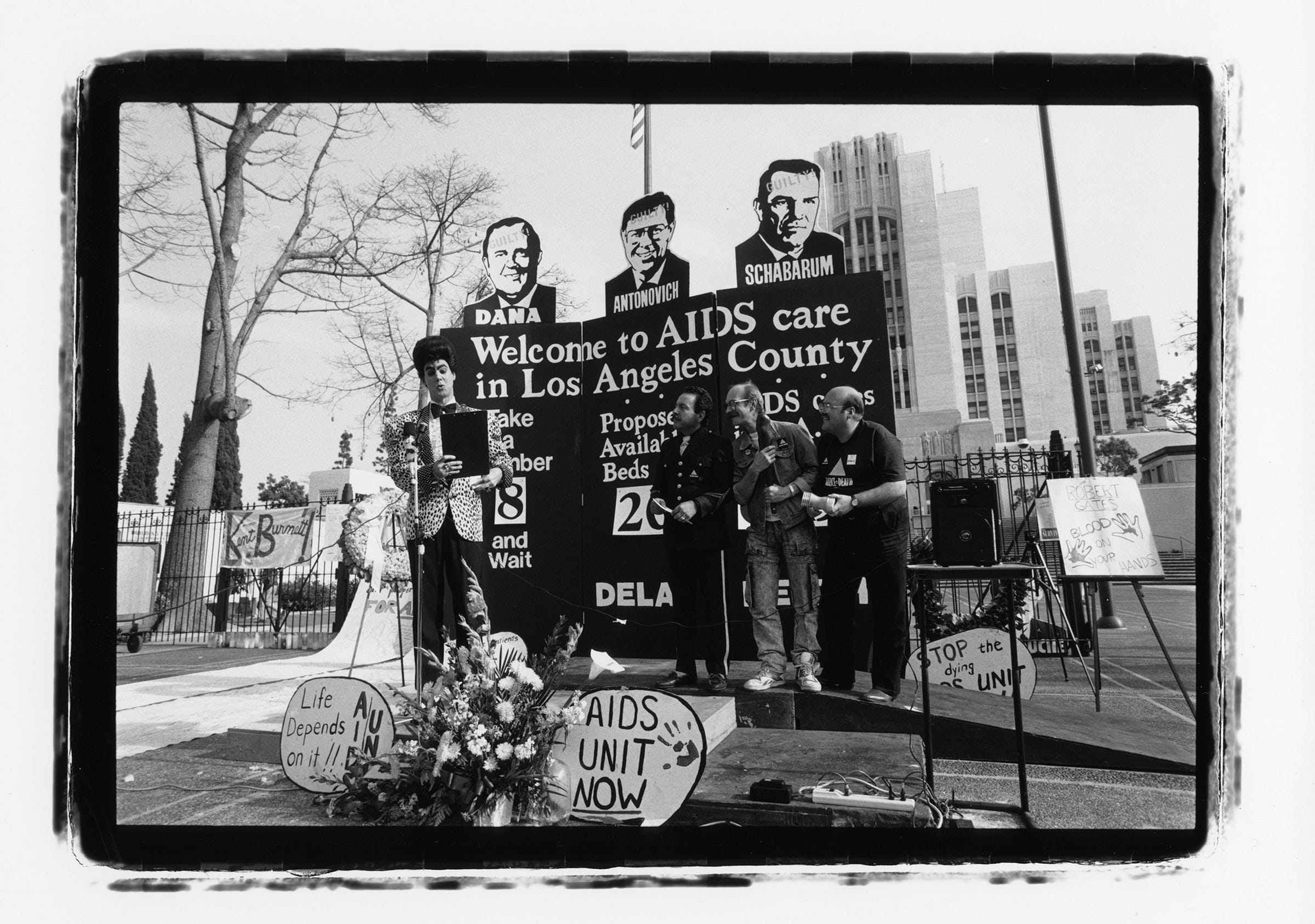 Activists hold a protest outside a hospital with a large backdrop explaining the AIDS crisis. "Welcome to AIDS care in LA County.. Take a number and wait... Proposed available beds 20... [Unreadable]. Signs saying AIDS UNIT NOW, Life Depends On It!! Some of the activists have dressed up like TV announcers or contestants. The backdrop lists several names. "Dana, Antonovich, Schabarum"