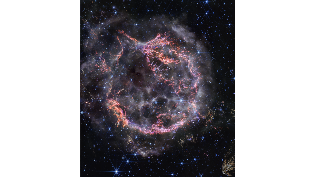 Cassiopeia A, a circular-shaped cloud of gas and dust with complex structure. The inner shell is made of bright pink and orange filaments studded with clumps and knots that look like tiny pieces of shattered glass. Around the exterior of the inner shell, particularly at the upper right, there are curtains of wispy gas that look like campfire smoke. The white smoke-like material also appears to fill the cavity of the inner shell, featuring structures shaped like large bubbles. Around and within the nebula, there are various stars seen as points of blue and white light. Outside the nebula, there are also clumps of yellow dust, with a particularly large clump at the bottom right corner that appears to have very detailed striations.