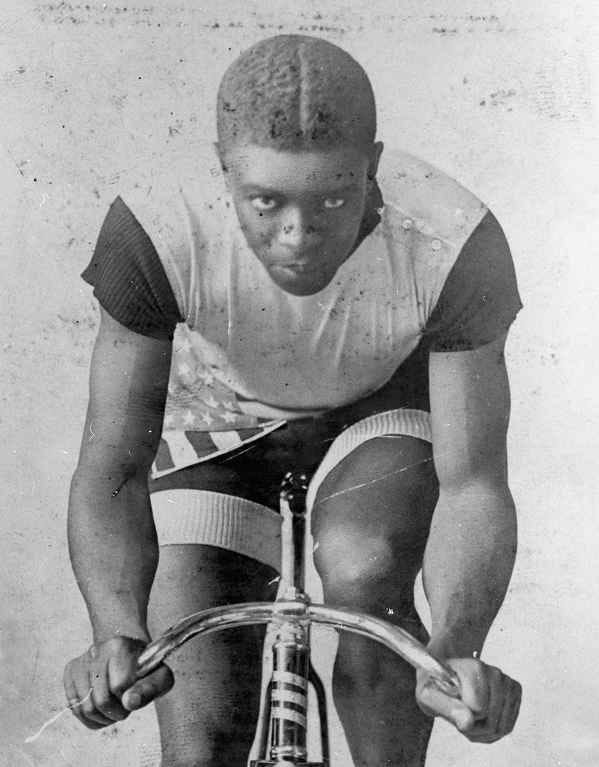 Old black and white photo of African American bicycle racer Major Taylor. Taylor is seated on a bicycle pointing directly at the camera, the feet on the pedals (left pedal higher, forcing his left knee higher than his right), his lean, muscular arms reaching forwards towards simple cowhorn shaped handlebars. His back is nearly flat and his head it tilted upward to directly face the camera. He is lean and muscular, his tightly trimmed hair parted neatly in the center. His face is young, his facial expression focused and serious. He is wearing a light-colored short sleeve top with dark, striped short sleeves. There are three buttons along the left shoulder. His cycling shorts are the reverse of the top, primarily dark with light, striped hems. Along his right hip is a stars and stripes sash of some sort.