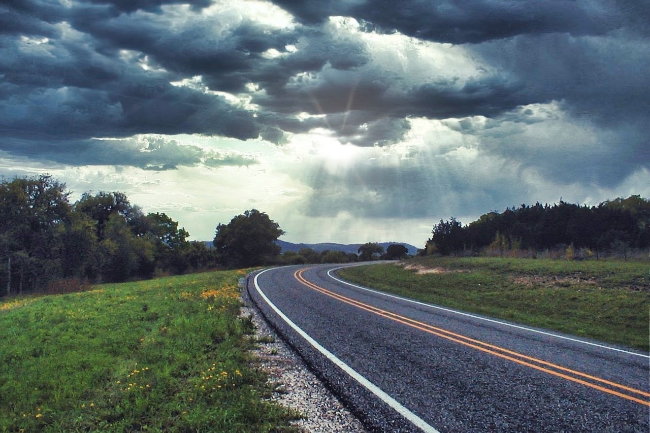 A view of a winding stretch of two-lane road with dark clouds in the sky and the sun streaming through