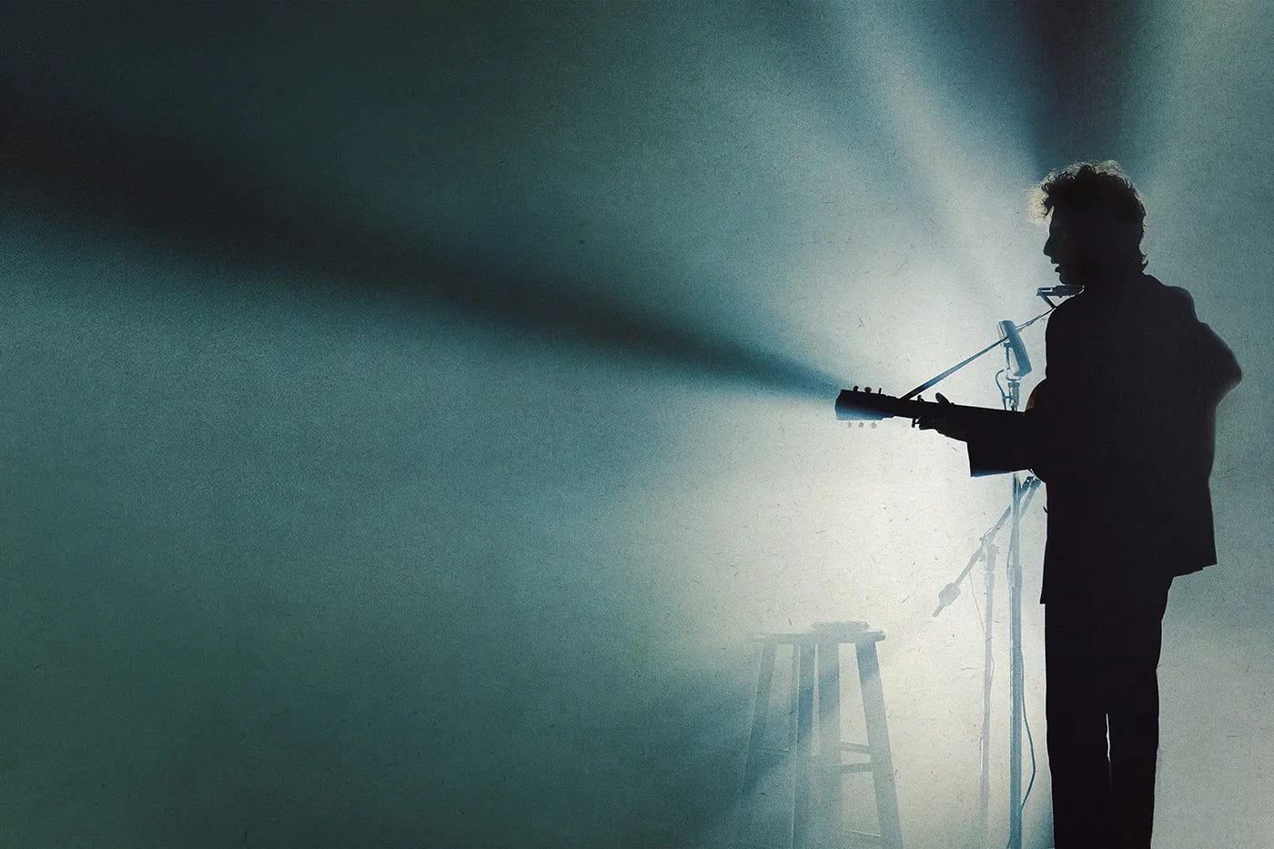 Timothée Chalamet holds a guitar onstage next to a mic stand and stool as Bob Dylan in the biopic A Great Unknown