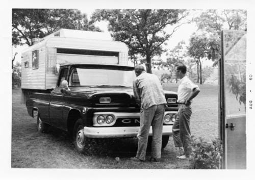 John Steinbeck, left, pops the hood of his camper van, Rocinante. His neighbor, Jack Ramsay (right), looks on. The black and white photo shows the camper van, build on a late 1950s Chevrolet pickup chassis, with a camper shell mounted to the rear.