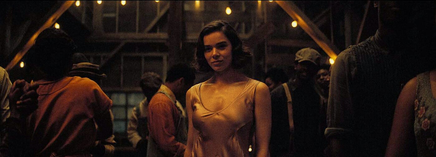 A woman in a satin slip stands centered among a crowd in a dimly lit barn dance, with warm string lights hanging overhead and faces watching her from the shadows.