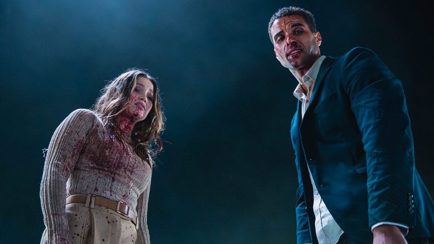 A blood-covered woman and a man in a torn suit look down at the camera, illuminated against a dark sky. A blood-covered woman and a man in a torn suit look down at the camera, illuminated against a dark sky.