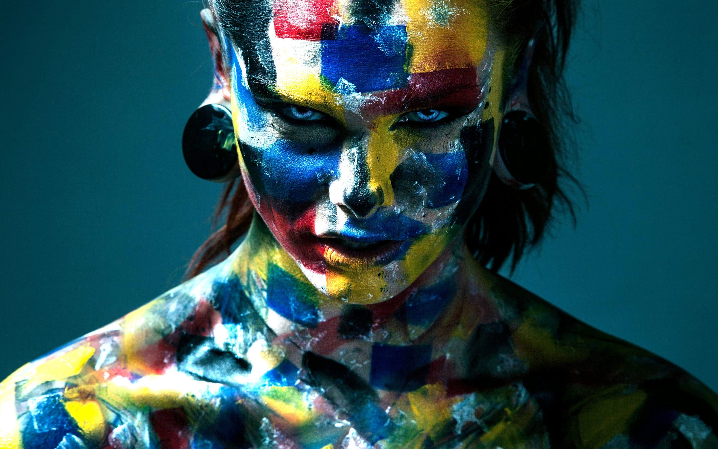 A man with icy blue eyes and square-shaped splotches of multicolored paint on his upper body and face stares intensely into the camera. The background is teal. A man with icy blue eyes and square-shaped splotches of multicolored paint on his upper body and face stares intensely into the camera. The background is teal.