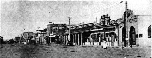 Black and white photo of a street in Elko, Nevada in 1910. The street is unpaved and there is a row of low one- and two-story structures facing the street. The taller buildings have advertising "Dry Goods" painted on the side of the upper floor. Some of the buildings have covered walks to provide shade. A row of telephone poles runs along the street.