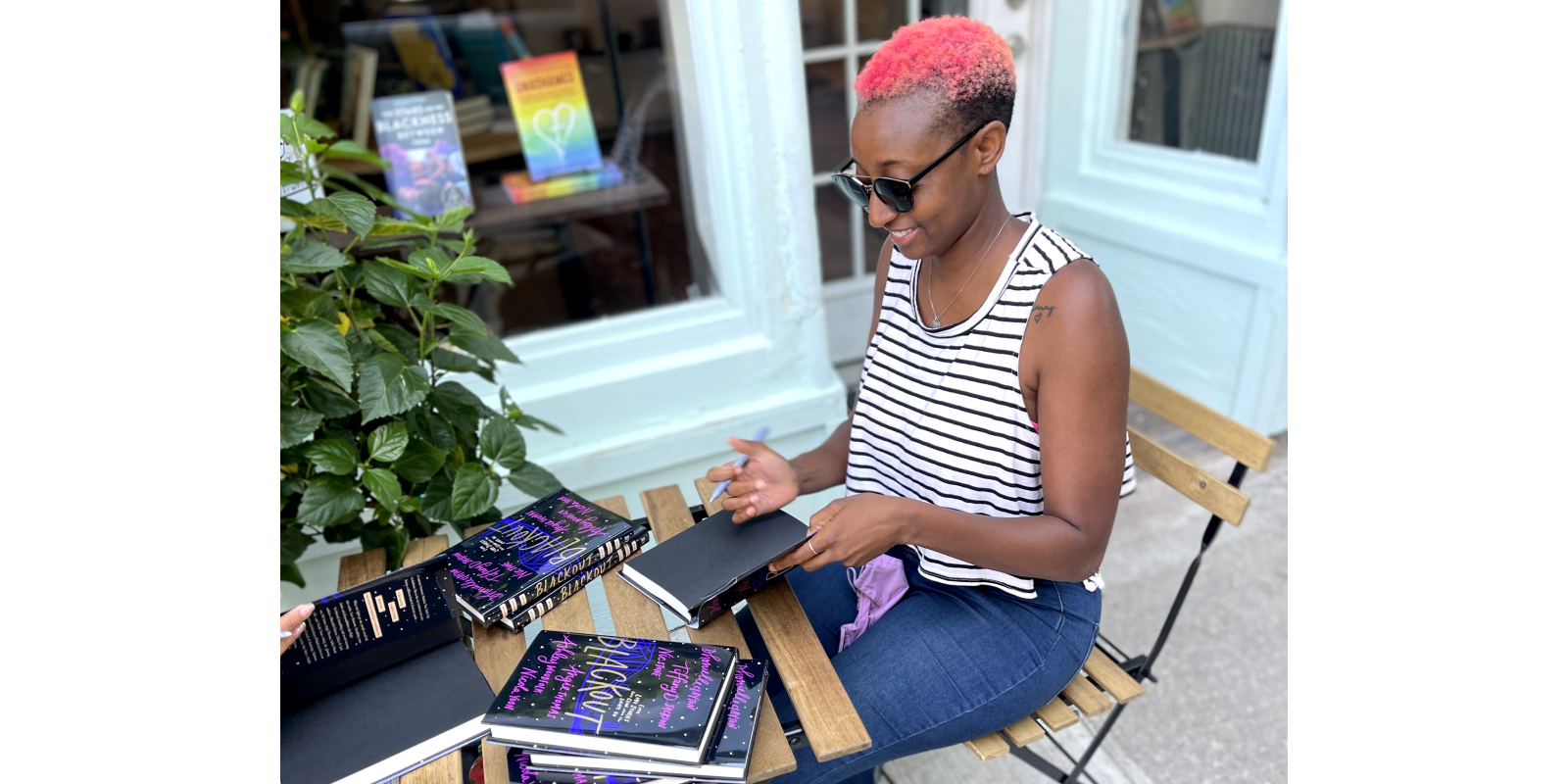 Ashley in pink hair and sunglasses, looking down and signing a book, outside a bookstore.