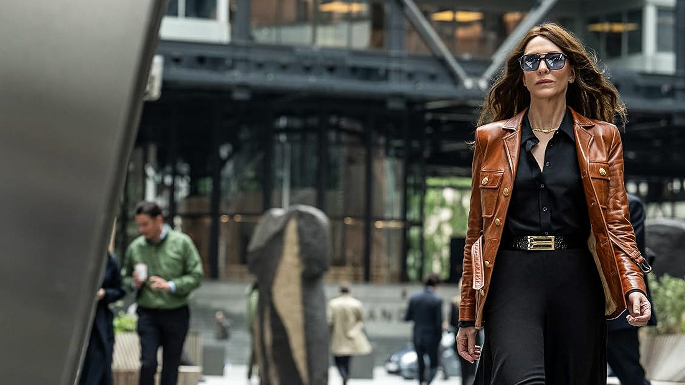 A woman in a rich brown leather jacket, black blouse, and slacks strides confidently through a modern office plaza. Her hair is wind-swept and sunglasses fixed. Blurred pedestrians move behind her.