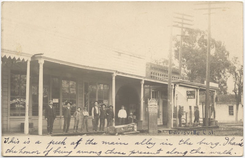 Old black and white photo of Davisville, California, featuring a row of simple one-story, brick and wood buildings with wooden walkways and awnings held up by simple columns. The building to the left features large glass windows upon with the word "Pool" can be seen. A row of men stand on the sidewalk and look at the camera. A wooden trough and pump (for horses) sits in front of another building, alongside a sign. Beyond, telephone poles rise. Below, handwritten, "This is a peek at the main ally in the burg. I have the honor of being among those present along the walk."