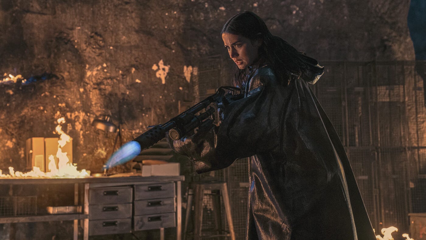 Ana de Armas as Eve Macarro, dressed in black tactical gear, aims a flamethrower inside an underground armory engulfed in fire.