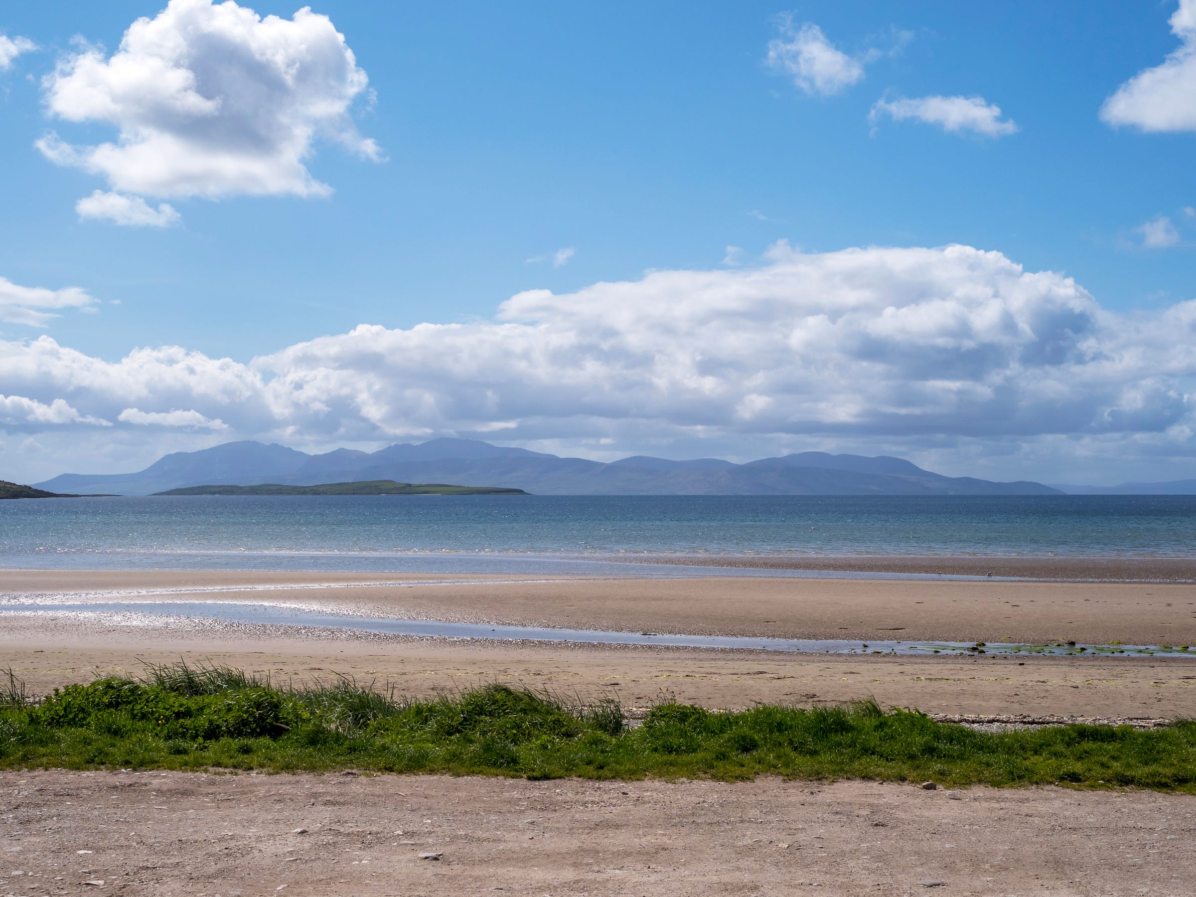 Photograph of a sandy beach at low tide on the Isle of Bute, Scotland. There is some green vegetation in the foreground, and a blue sky above with white clouds.  There is a view over the sea to the Isle of Arran.