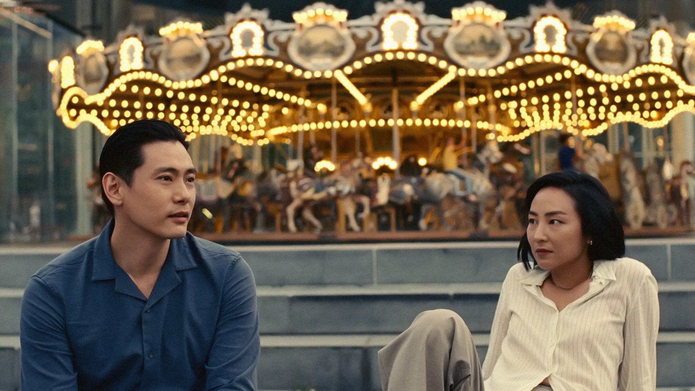 A man and woman sitting in front of a carousel. He is looking off in the distance and she is leaning back and looking at him.