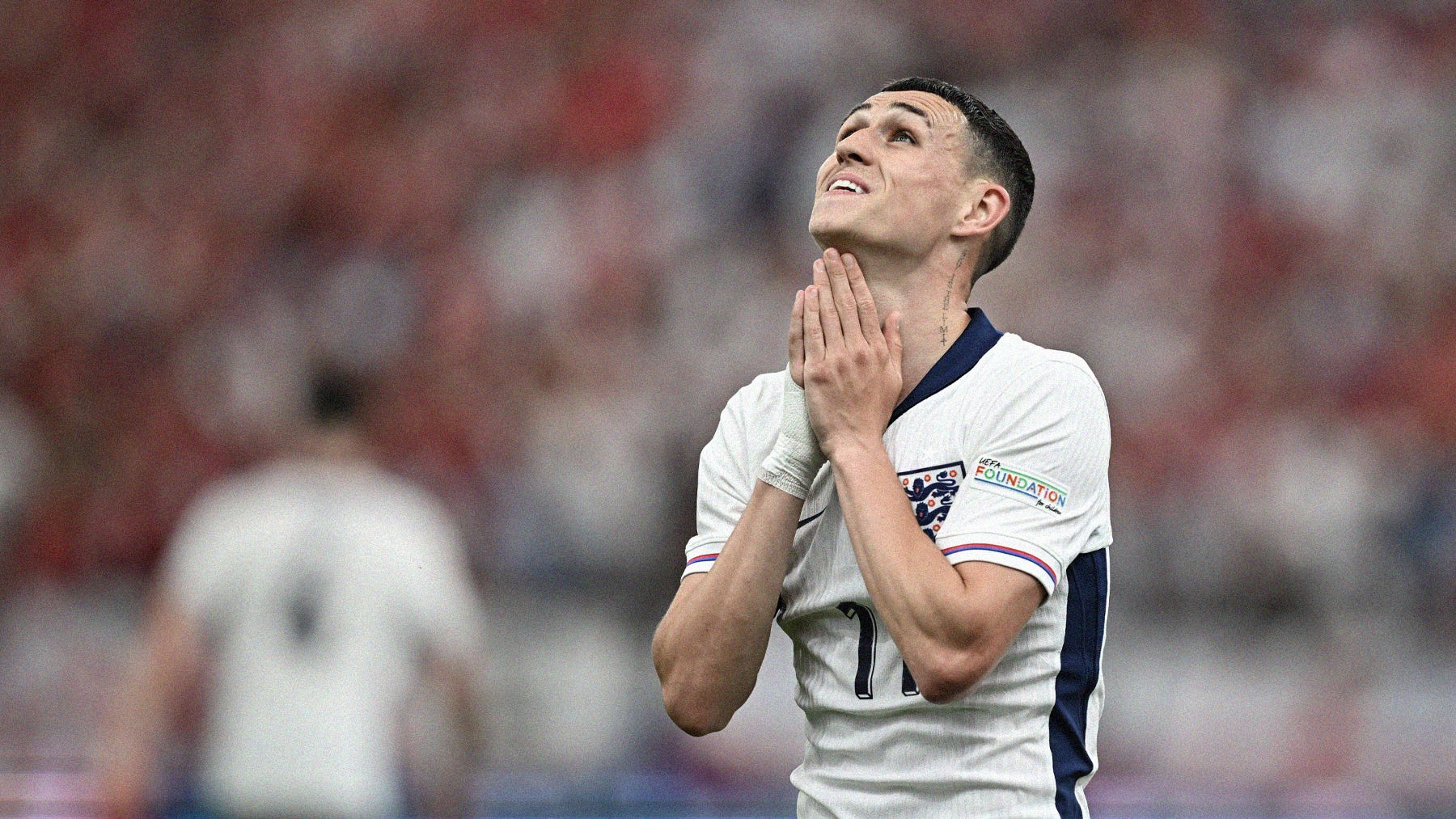 A photo of Phil Foden looking up to the sky with his hands in a praying position. He's wearing a white England shirt with navy trim