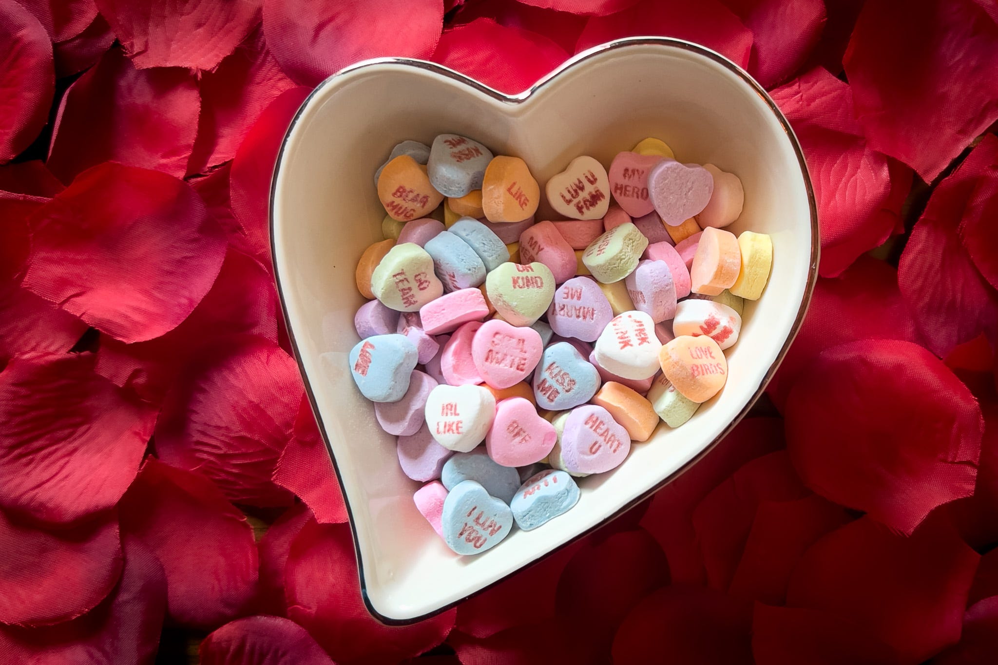 A heart shaped bowl sitting in a bed of  red rose petals with small candy hearts inscribed messages, like “be kind” and “love u”