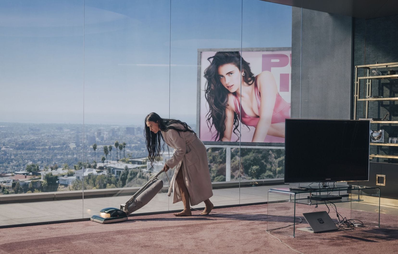 Demi Moore as Elisabeth Sparkle in the film The Substance vacuums a carpet in front of a large glass window. From the window, you can see a giant billboard for a new television show starring the much younger Sue, played by Margaret Qualley.