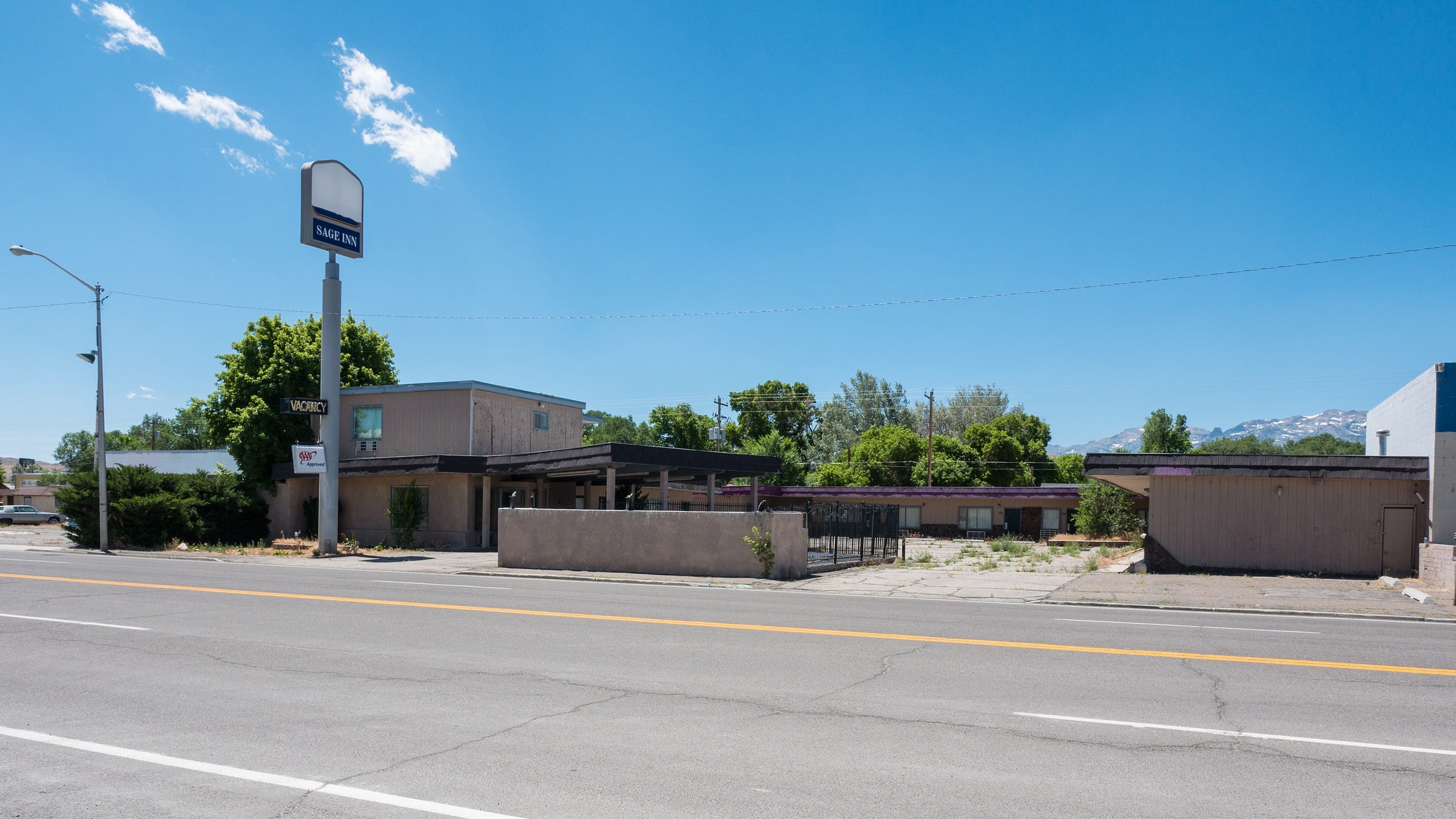 Photo looking across an empty street at an abandoned motel. It is a bright, sunny day with just a handful of clouds dotting the sky. A tall, 30 foot pole stands on the curb with the motel's old lighted sign at the top, but the name has been removed. A small, empty pool is near the entrance, surrounded by a simple black metal fence. The motel is u-shaped with a central courtyard/parking area. Tall weeds grow through cracks in the pavement. Trees grow behind the motel and jagged mountains can be seen in the distance.