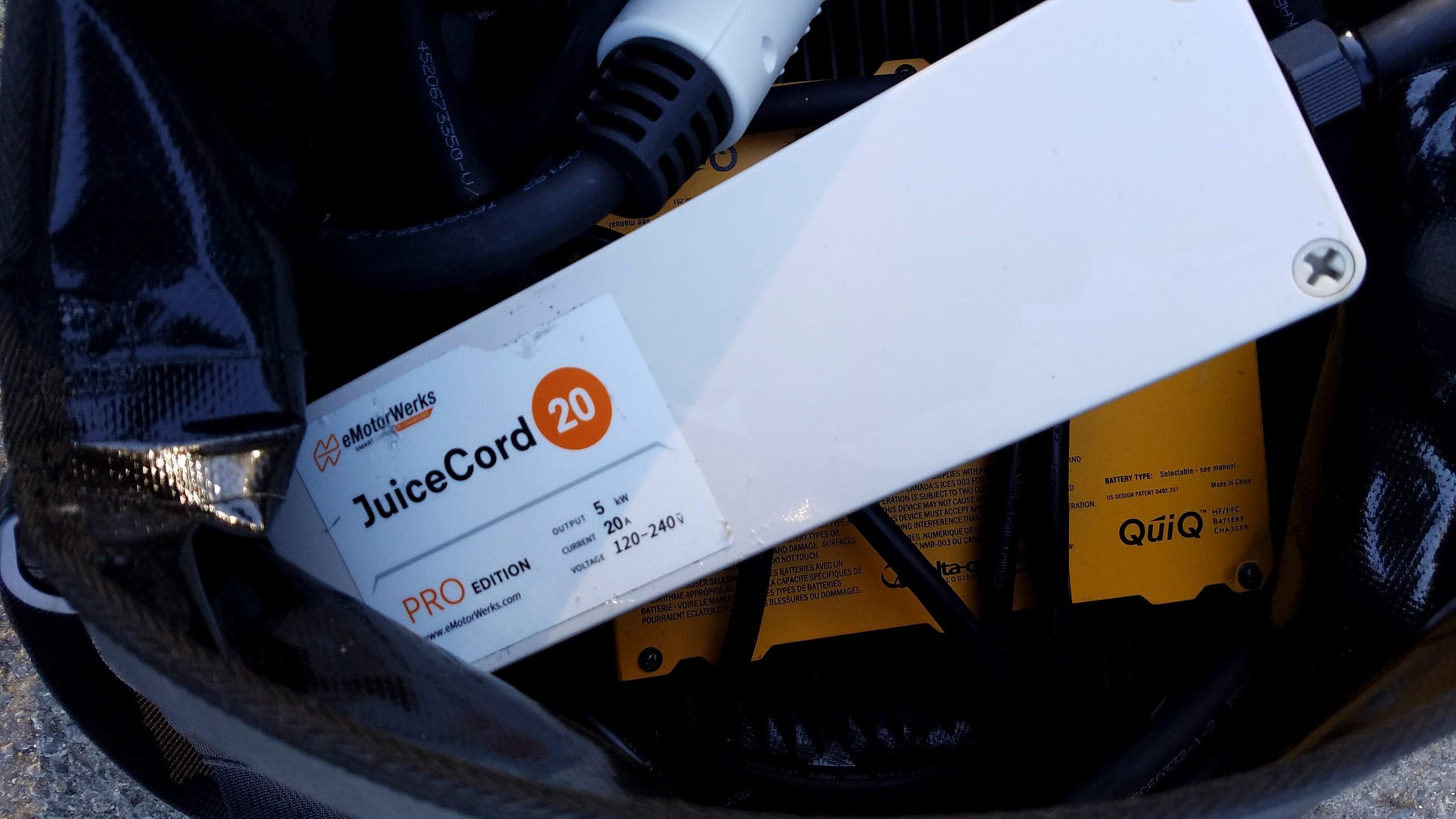 Closeup photo of a black waterproof duffle bag. The bag is open, revealing an orange box with the label "QuiQ" in the bottom of the bag and a white box with the label "eMotorWerks JuiceCord 20 PRO Edition". At the top of the photo is a white connector for an EV charging station.
