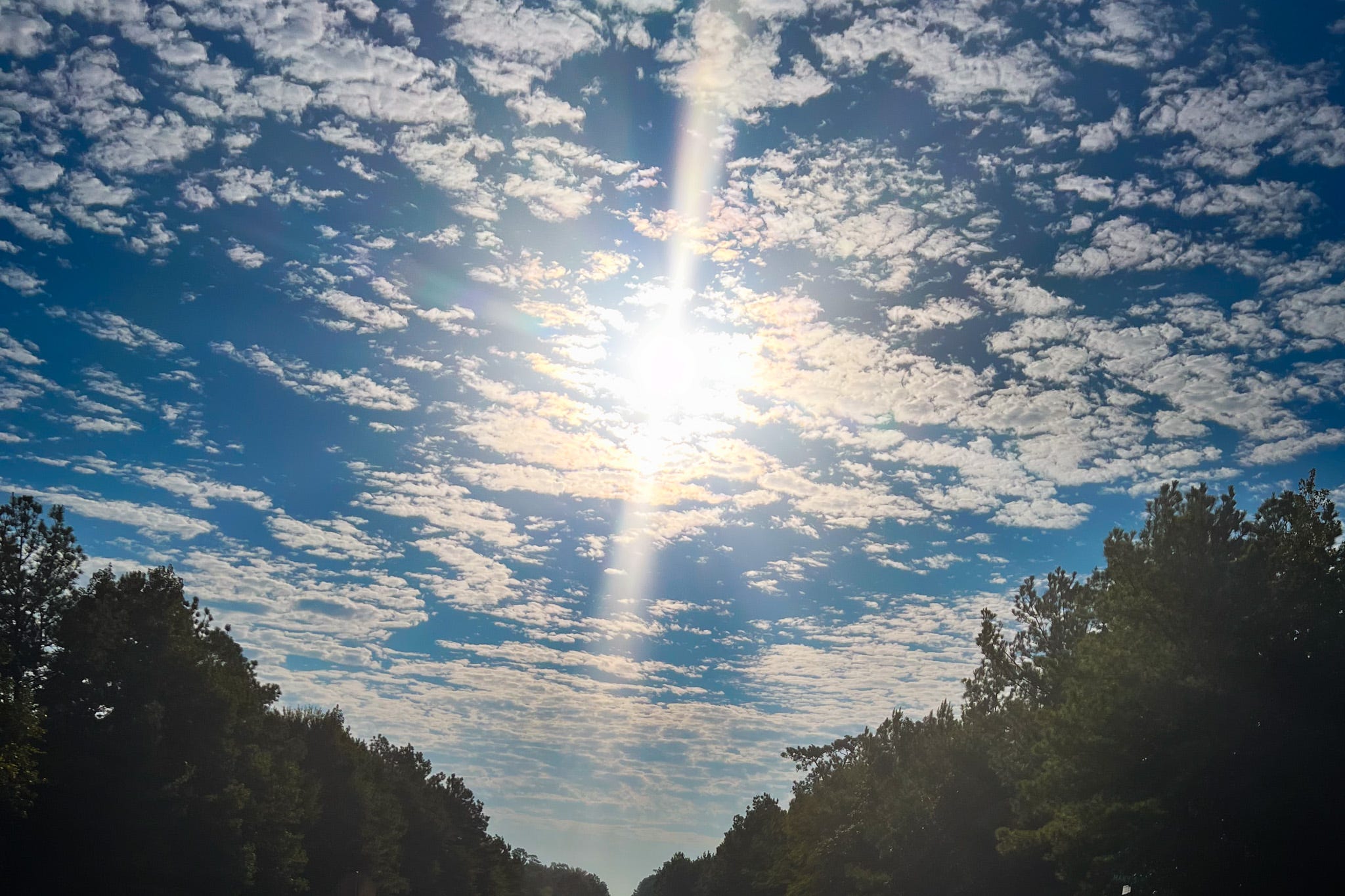 The mid-day sun shining brightly behind a scattering of clouds with a blue sky in the background. Trees create two triangles and both sides of the photo