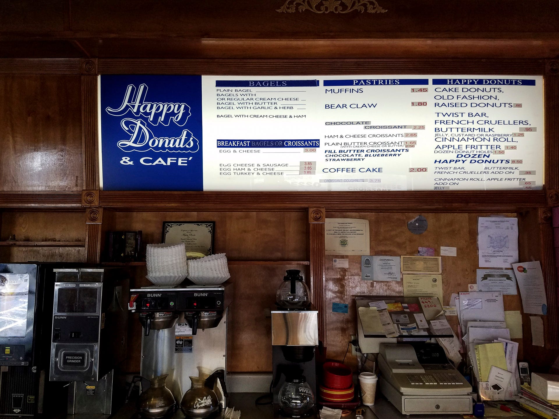 Photo of the back wall of "Happy Donuts & Cafe". On the left side of counter sit three industrial-sized Bunn coffee makers, a coffee grinder, and a cream dispenser. On the right side is a cash register, various legal certificates affixed to the wall, and various and sundry papers, envelopes and detritus. The back wall is finished in stained wood, with shelves and trim pieces that have seen better days. Above is the backlit "Happy Donuts Cafe", blue lettering and graphics upon a white background. Categories are "BAGELS", "BREAKFAST BAGELS OR CROISSANTS", "PASTRIES", and "HAPPY DONUTS" with items listed with each category. Prices are printed on white paper (in red lettering) and then affixed beside each item. Lettering seems haphazard.