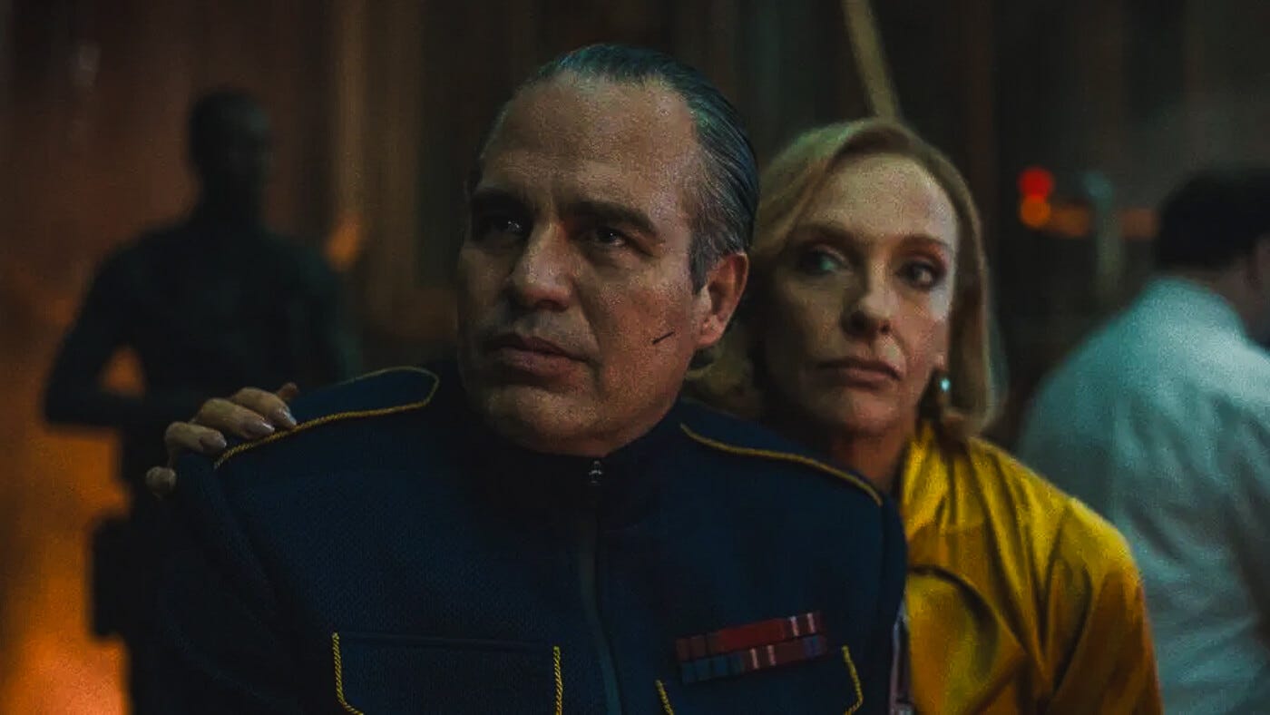 Mark Ruffalo in a navy authoritarian uniform looks ahead sternly while Toni Collette, in a bright yellow coat, stands behind him with a skeptical expression.