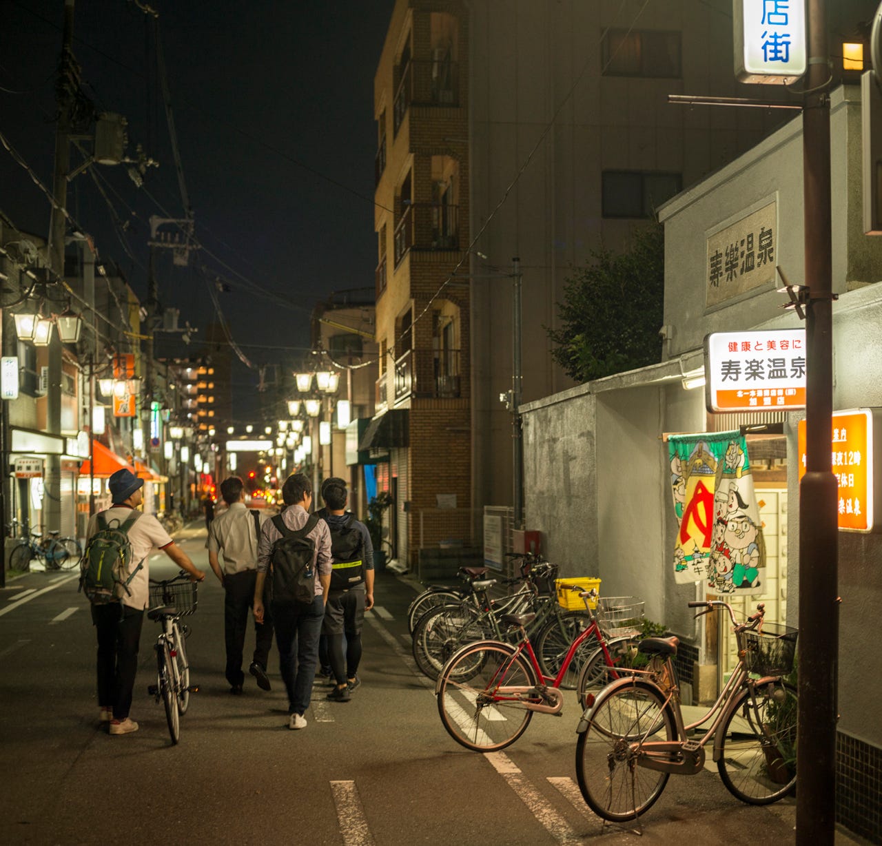 A group of young men walk by Juraku, an old community bath house, still lit up and in use in Kitakagaya, Osaka, Japan