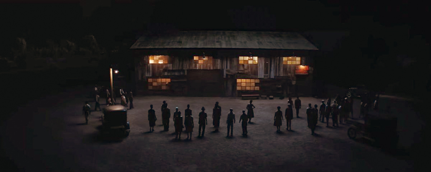 A wide shot of a barn at night with silhouetted townspeople standing before it, their shadows stretching toward the camera under a patchwork of warm, interior lights.