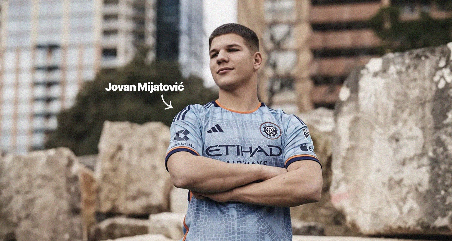 A photo of Jovan MIjatovic stood with his arms crossed, looking away from the camera with a smile on his face, wearing a sky blue NYCFC shirt.