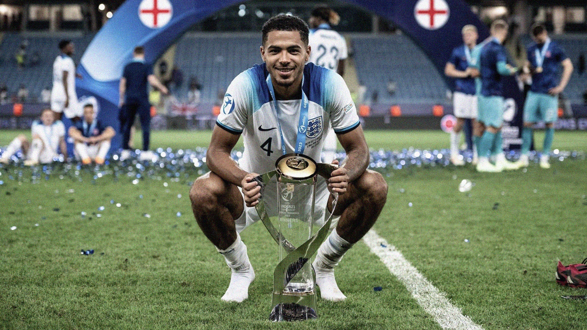 A photo of England's Levi Colwill crouching down, holding the UEFA U-21 EURO by either side, smiling at the camera