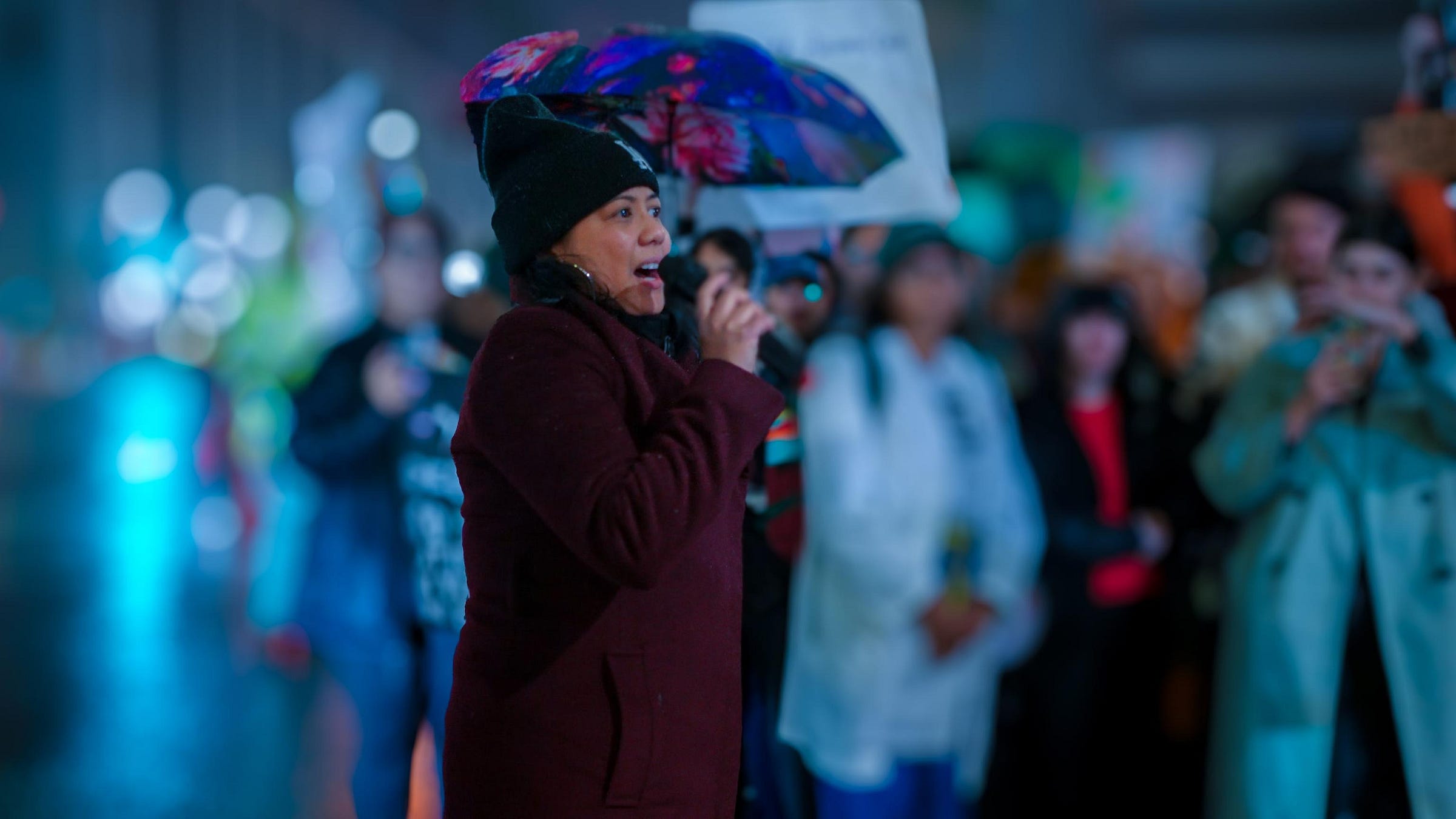 Ysabel Jurado holds a microphone surrounded by a blurred crowd of protestors with umbrellas and signs