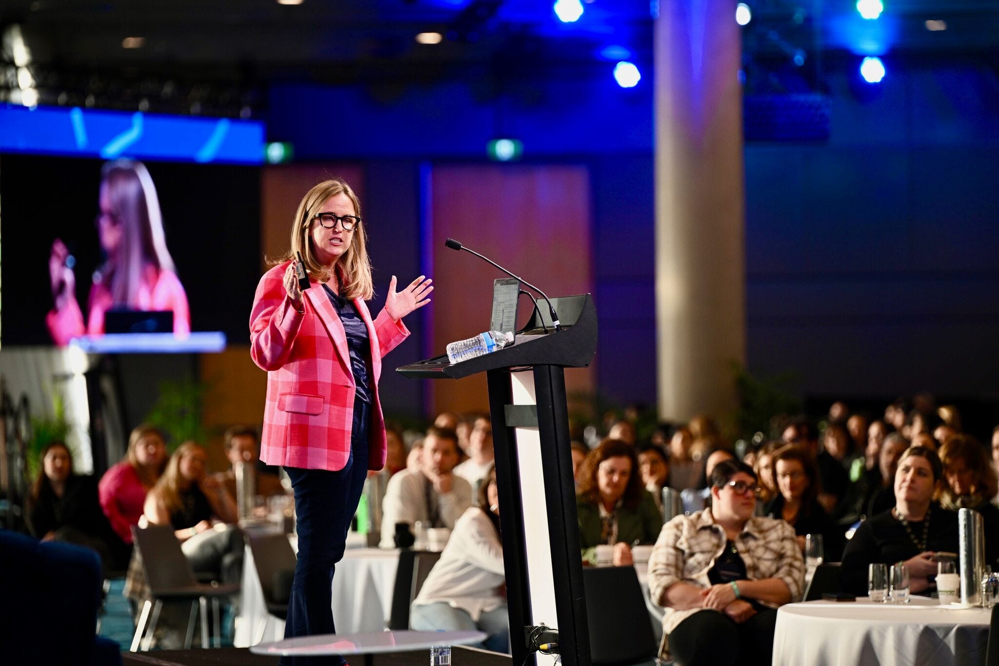 Lia Haberman standing at a podium in front of a blurry audience speaking at the State of Social conference wearing an amazing red and pink jacket from Trina Turk that manifests good fortune xo Lia Haberman standing at a podium in front of a blurry audience speaking at the State of Social conference wearing an amazing red and pink jacket from Trina Turk that manifests good fortune xo