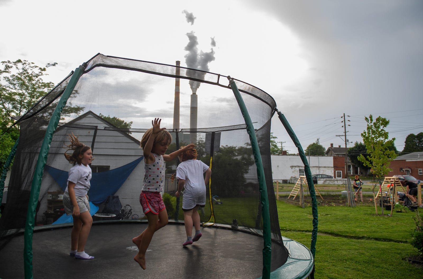 Three children jump on a trampoline outside on a cloudy day. Behind them is a white house and a tall smokestack emitting grey smoke.