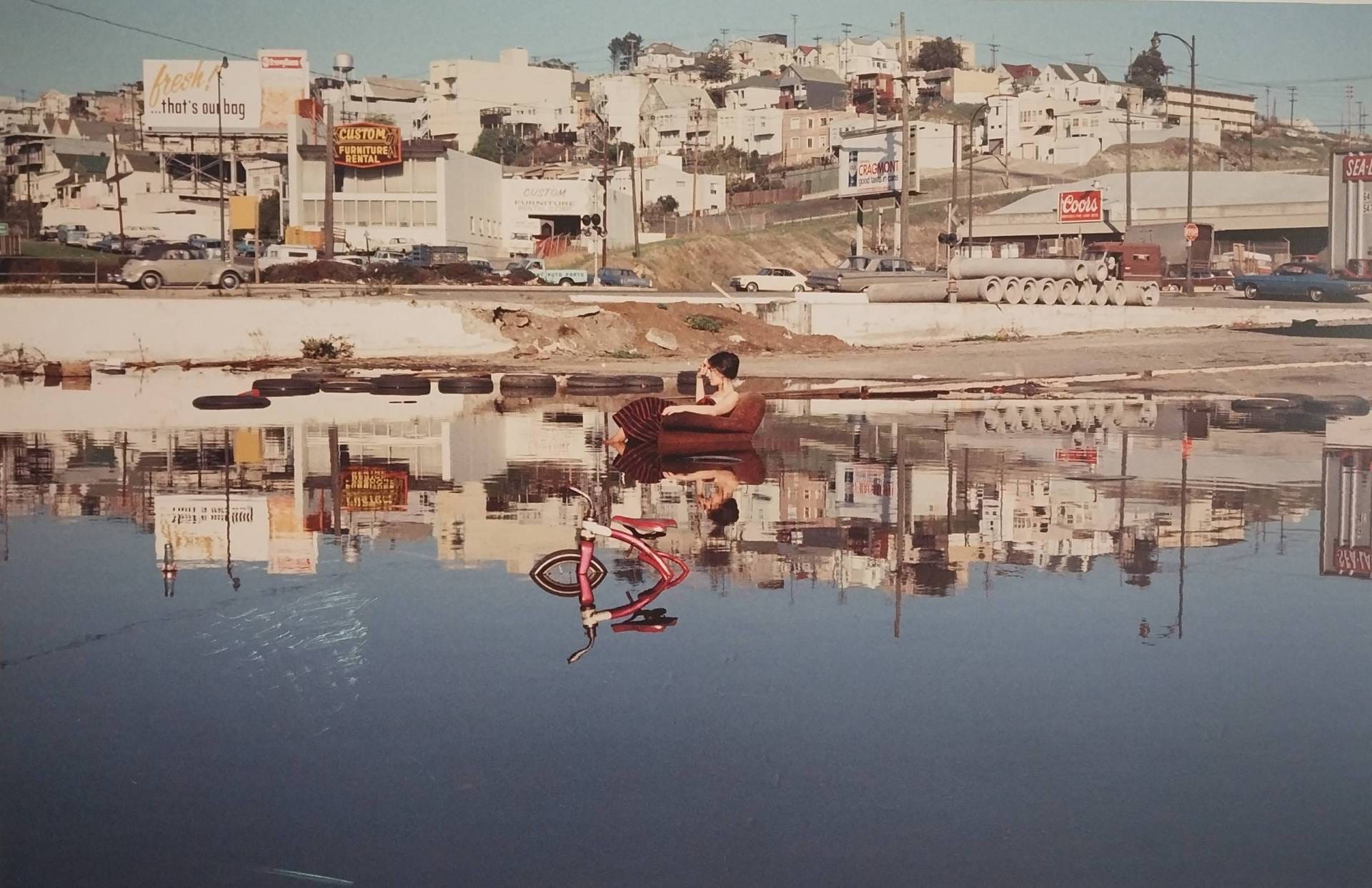 A woman casually sits in an armchair smoking a cigarette in the middle of a flooded parking lot. Urban houses are seen behind her.