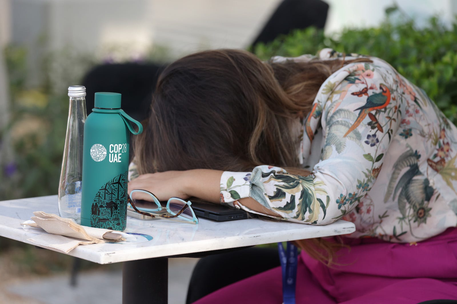 A woman lays her head on a faux marble desk during a break in COP28 for a nap. Only her brown hair, glasses, and green COP28 UAE water bottle can be seen.