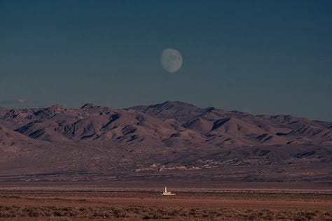 Three images from Postcards From the End of the World, taken by Victoria Scott. First is a desert landscape at dawn with a mountain range in the background and the moon looming large over them; second is a tower of some kind, looking deserted and aglow by pinkish light in the darkness of dusk; third is an old-time outpost situated on the banks of a river fed by mountains in the background green and flush from recent rains.