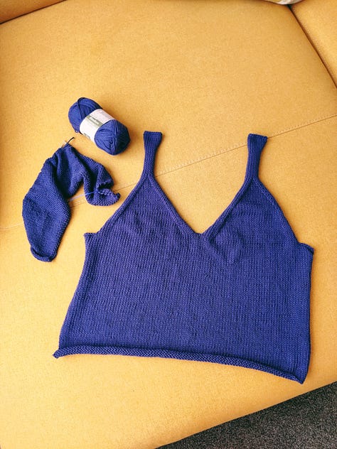 A series of craft projects: 1) Half a knitted singlet in bright colbalt blue, with a v-neck and the other half started on needles laying nearby. 2) A selfie of Louise wearing a matching pair of pyjamas with big yellow pears on them. 3) A selfie of Louise wearing a knitted singlet with a round-neck, in shades of grey, black, and white. 4) Two pairs of trouser legs laid over the top of each other, one is pale blue and the hem has been unpicked, one is deep brown-pink and is much shorter. 5) A close-up of some mending of a jumper with large tomatoes on it. 6) A seflie of Louise wearing the tomato jumper. 7) Two matching knitted hats, one in shades of green, one in shades of pink, both have '2022' labels on them. 8) A pale pink cropped blouse with white abstract faces, and a v-neck and ruffle sleeves on a coat hanger. 9) A photograph of a folded linen scarf, it is gingham with shades of white and beige. 