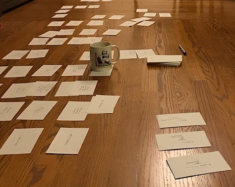 Index cards with writing on them, arranged on a hardwood floor with a mug of tea that reads "Book Nerd." Two large piece of white butcher paper with a color-coded outline, on the floor with other pages and a laptop. A piece of white butcher paper covered in writing, unfolded on a table. 