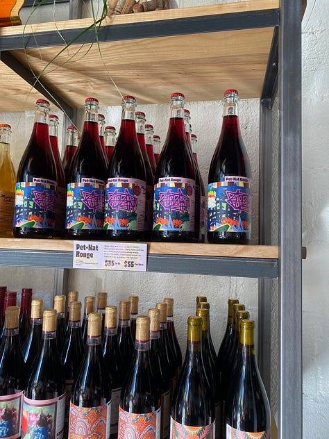 Wine bottles with images of my artwork on the labels arranged in rows on shelves. In the middle is a photo of me, a man holding a bottle of the Pet-Nat Rouge smiling for the photo and looking out at the viewer.